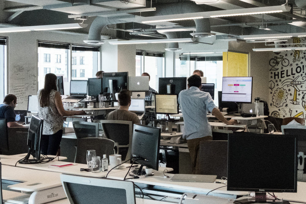 Open-plan technology company office with employee workstations and a whiteboard visible in the background